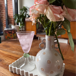 Wide circular rim pink glass glistening with evanescence bubbles. It narrows into a circular green glass, sitting atop a clear glass stand. The cocktail glass sits perfectly on a wooden table next to a polka dot vase holding a beautiful array of pink roses